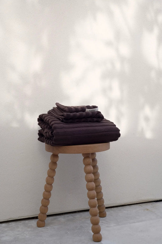 Stacked Chocolate home towel set on a timber stool in dappled light