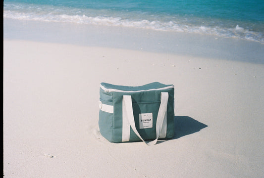 Tallow soft cooler bag resting on the sand beside clear blue water